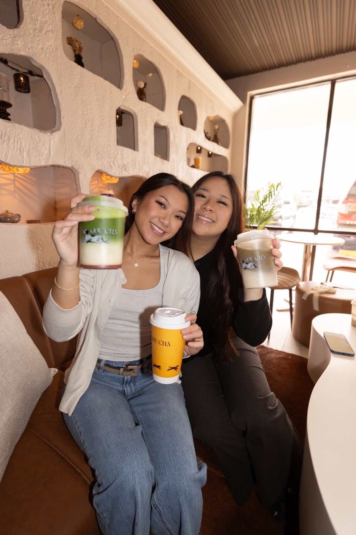 Sisters Kim Vo and Tammy Lien smiling and holding Oou Cha drinks on the café's leather couch