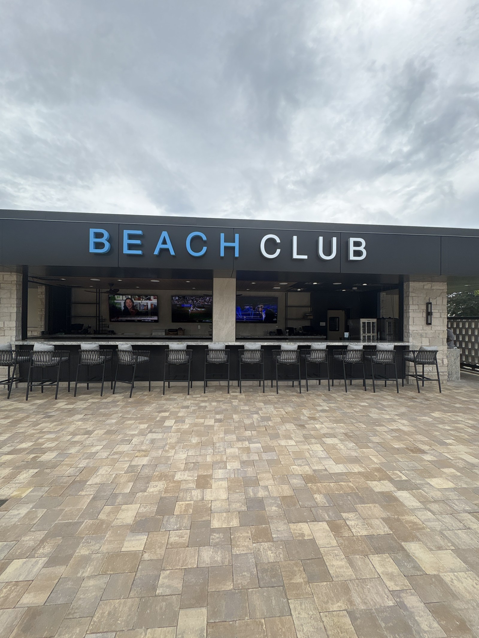 An outdoor bar with stools and TVs under a Beach Club sign.