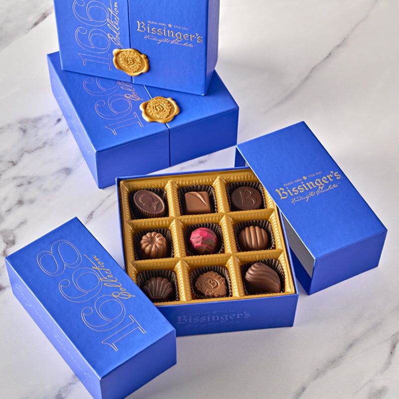 An open blue Bissinger's 1668 Collection box showing nine chocolates in a gold tray, beside closed boxes with gold wax seals.